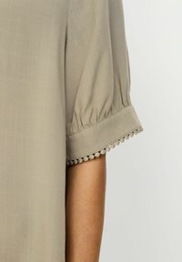 Svijetlo siva bluza s napuhanim kratkim rukavima, koja ima nabornu manžetnu i delikatni pom-pom ukras duž rubova, izrađena od mekanog, teksturiranog materijala.