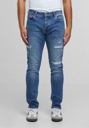 Kék farmernadrág, slim fit szabással, kopott részletekkel, szakításokkal és festékszórásos díszítéssel. Fehér edzőcipővel viselve.