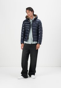 Marineblauwe pufferjack met een rits aan de voorkant en een capuchon, met een gestikt ontwerp. Gekozen boven een lichtblauwe shirt, gecombineerd met donkerblauwe jeans en witte sneakers.