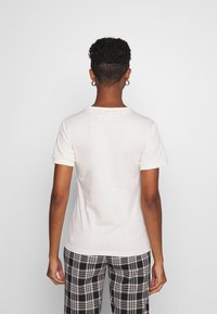 Krémové bavlněné tričko s ohrnutými rukávy; zadní pohled ukazuje hladký, přiléhavý střih. V kombinaci s černobílými kostkovanými kalhotami.