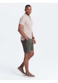 Beige polo triko spojené s tmavě zelenými kraťasy, vyrobené z měkkého materiálu. Doplňují je světle hnědé pantofle a náramkové hodinky.