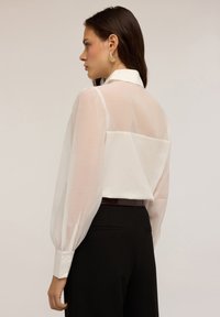 Femme avec des cheveux foncés portant un blouse en voile blanc à manches longues rentrée dans un pantalon noir taille haute, de profil contre un fond uni.