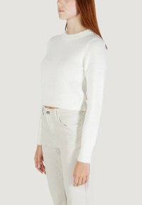 Una donna con capelli rossi lisci indossa un maglione cropped bianco e peloso con maniche lunghe e pantaloni beige chiaro a vita alta su uno sfondo semplice.
