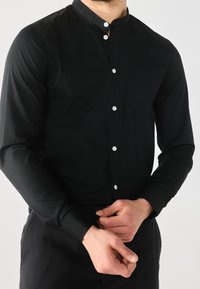 Zwart shirt met lange mouwen en een opstaande kraag, voorzien van zilveren knopen en een borstzak. Soepele textuur en aansluitend ontwerp.