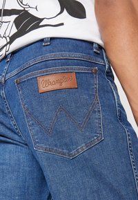 Krupni plan plave Wrangler traperice s stražnjim džepom, smešnim kožnim logotipom i narančastim šavovima, nošene s bijelom majicom.