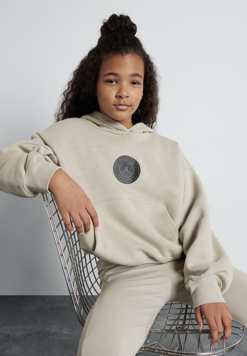 Felpa con cappuccio beige con una grande patch circolare che presenta il testo "PARIS". Texture liscia, vestibilità oversize e polsini a costine.