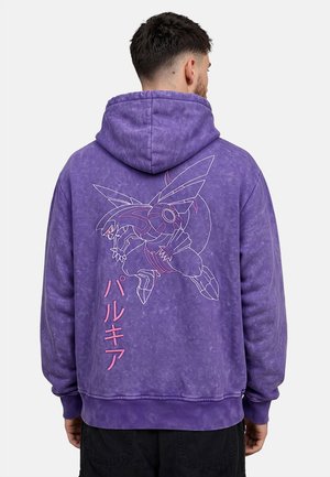 Uomo che indossa una felpa con cappuccio viola con un grande disegno stilizzato di un insetto e testo giapponese sulla schiena, in piedi su uno sfondo semplice.