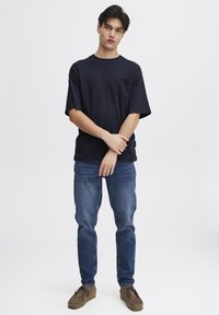 Tricou texturat bleumarin cu buzunar pe piept, asortat cu blugi bleumarin și pantofi din suede maro. Modelul stă în picioare pe un fundal alb.