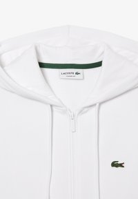 Sweat à capuche blanc zippé avec cordons, présentant un logo crocodile brodé vert et rouge sur la poitrine et une étiquette "Lacoste Classic Fit" à l'intérieur du col.