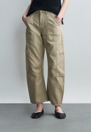 Személy laza, bézs színű, több zsebbel rendelkező cargo nadrágban, fekete lapos cipőkkel, kezét a zsebében tartva áll.