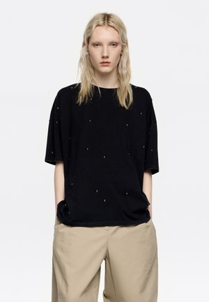 Camiseta negra de manga corta con un ajuste holgado, adornada con tachuelas metálicas dispersas. Combinada con pantalones beis, que presentan un diseño simple y minimalista.
