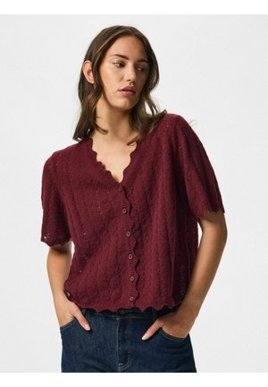 Kvinde iført en vinrød strikket kortærmet knap-cardigan med bølgede kanter, kombineret med mørkeblå denimjeans, hænderne i lommerne.