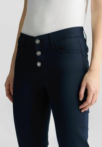 Vaqueros de mezclilla azul marino con cintura alta, que presentan un cierre de cinco botones, bolsillos y detalles de costura sutiles. Textura suave y diseño ajustado.