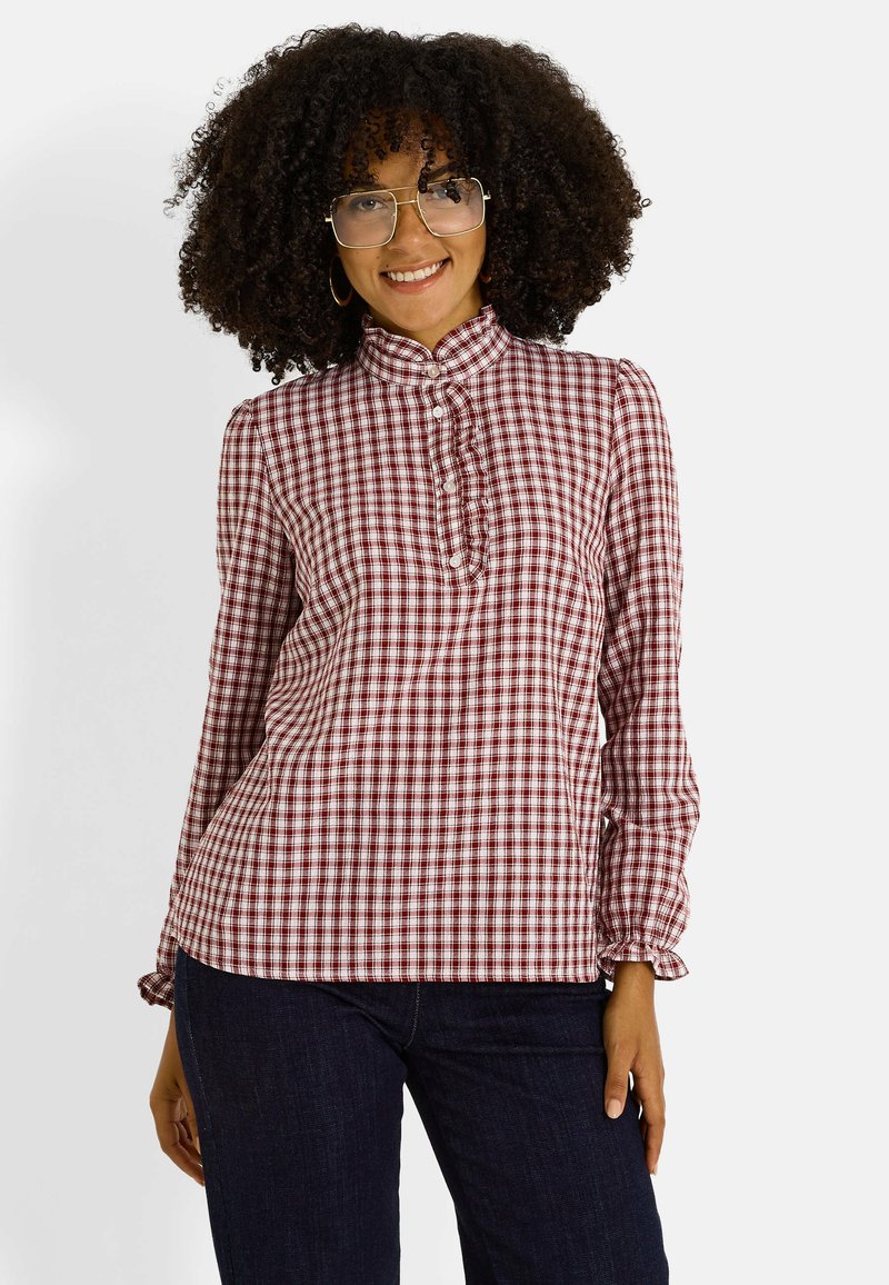 Blusa a cuadros rojos y blancos con cuello alto, tapeta de botones y detalles de volantes. Mangas largas con puños elásticos. Llevada con jeans oscuros.