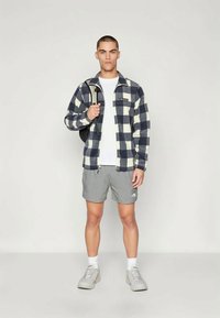 Herre flanelljakke i marineblå og kremfarget rutemønster, grå shorts og hvit T-skjorte, kombinert med grå sko. Ryggsekk over den ene skulderen.