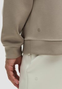 Beige sweatshirt met ribbelboorden en -zoom, voorzien van een klein, reliëf logo. Soepele textuur, gecombineerd met lichtgekleurde onderkleding.