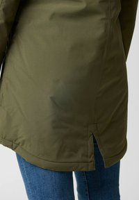 Olijfgroene parka met een gladde textuur, voorzien van zijsplitten aan de zoom en gestikte details. Draagt over blauwe spijkerbroek.