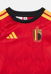 Κόκκινη φανέλα με μοτίβο φλόγας, μαύρο λαιμό με ραφή και κίτρινα διακοσμητικά στοιχεία. Διαθέτει κεντημένο λογότυπο της Βασιλικής Ομοσπονδίας Ποδοσφαίρου του Βελγίου.