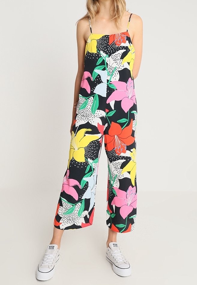 Femme portant une combinaison noire à jambes larges avec un grand imprimé floral coloré et fines bretelles, associée à des baskets blanches basses.