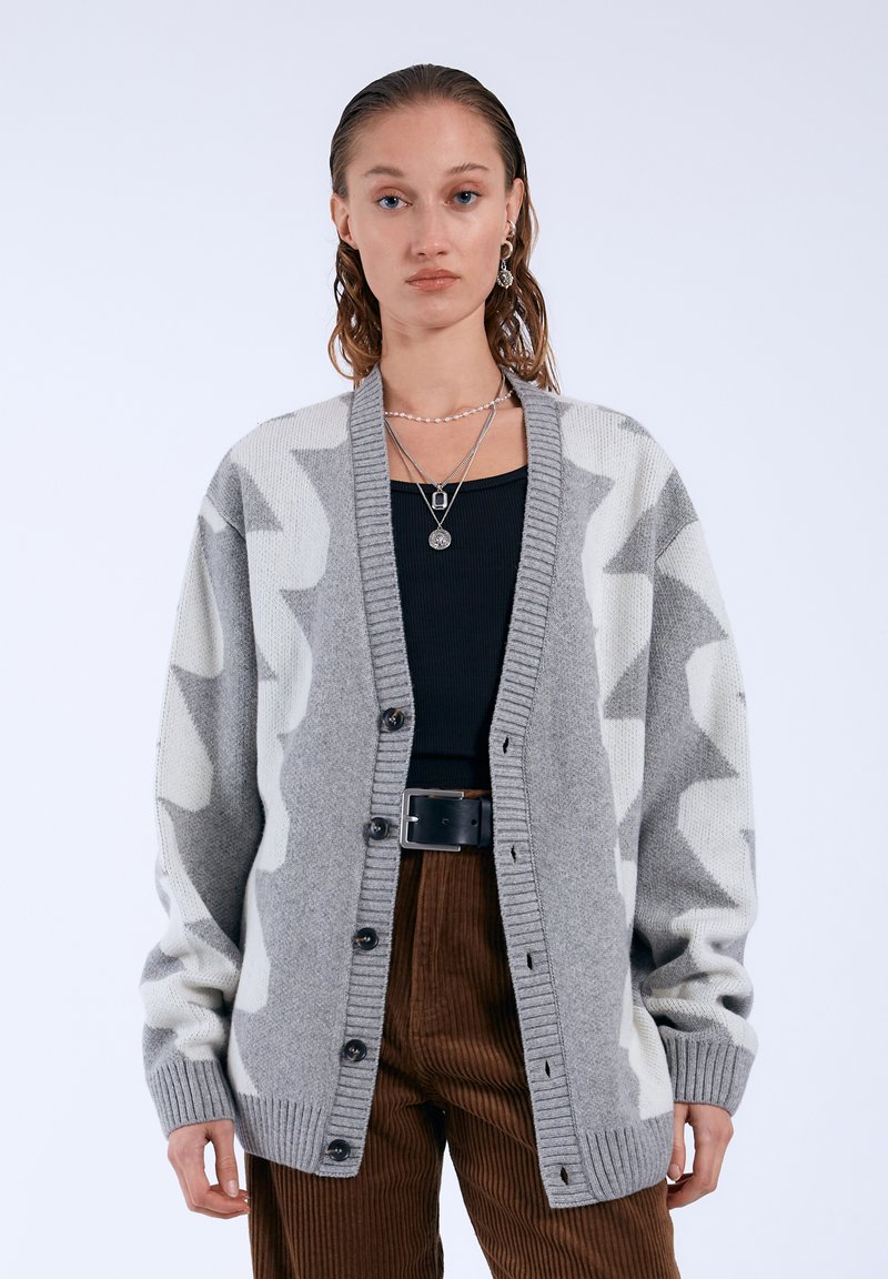 Gray knit cardigan featuring a white zigzag pattern, button closure, ribbed cuffs, and a relaxed fit, paired with a black top and brown corduroy pants.