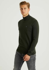 Suéter de cuello alto verde oscuro con un corte ajustado. Presenta un cuello y puños de canalé, textura lisa y un pequeño logo negro en el pecho.