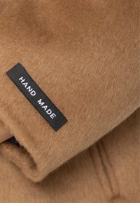 Beige blødt uldstof med en sort label syet på, der viser teksten "HAND MADE" med hvide store bogstaver.