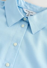Camicia Oxford azzurro chiaro realizzata in tessuto testurizzato, caratterizzata da un colletto a punta, una pattina con bottoni bianchi e dettagli di cucitura visibili.