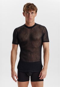 Sort mesh kortærmet skjorte med en rund halsudskæring, der har en løs pasform og åbent strikmønster. Bæres over tætsiddende sorte shorts.