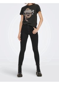 Camiseta gráfica negra con un diseño de serpiente y texto, combinada con jeans ajustados negros y botas de combate negras, todo en un atuendo casual.