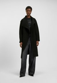 Długi czarny płaszcz o teksturowanej powierzchni z szerokim kołnierzem i dwoma guzikami, zestawiony z ciemnymi spodniami o szerokich nogawkach, z delikatnym wzorem.