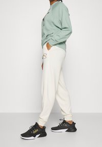 Light green sweatshirt with ribbed cuffs, cream joggers with a circular logo, and black sneakers with gold accents and white soles.