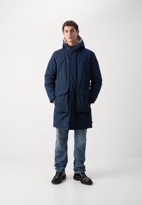 Abrigo aislante azul marino con capucha, que presenta dos bolsillos frontales, cierre de botones y un interior texturizado, combinado con jeans de color azul claro y zapatos negros.