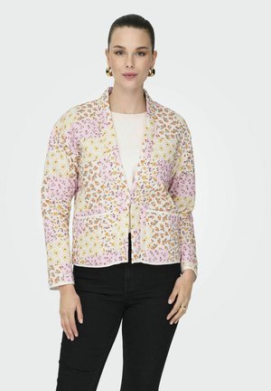 Femme portant une veste patchwork florale aux tons rose, blanc et orange, sur un haut blanc et un pantalon noir, sur un fond uni.