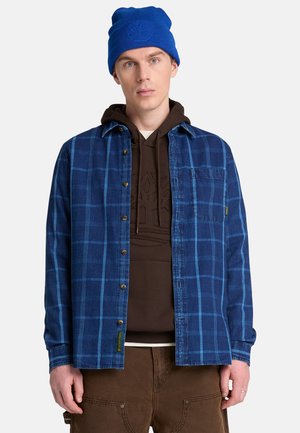Mann trägt eine blaue Mütze, ein blau kariertes Hemd über einem braunen Kapuzenpullover und braune Hose, steht vor einem einfarbigen hellen Hintergrund.
