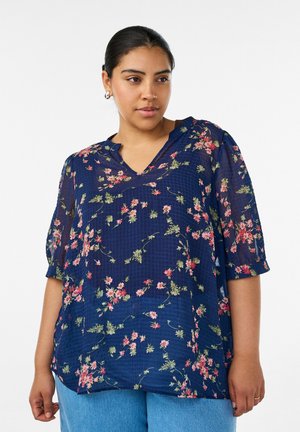 Kvinde iført en marineblå blomstret bluse med trekvartlange ærmer og lyseblå jeans, stående mod en hvid baggrund.