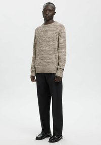 Beiger Strickpullover mit strukturiertem Muster, rundem Ausschnitt und gerippten Bündchen, kombiniert mit schwarzen Hose und schwarzen Loafers.
