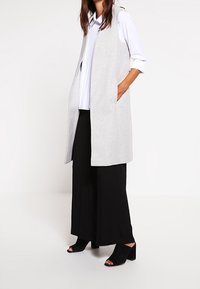 Grå ärmlös väst med sidofickor, över en vit plisserad skjorta, i kombination med svarta vida byxor och öppna svarta klackar.