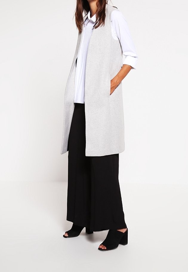 Grå ärmlös väst med sidofickor, över en vit plisserad skjorta, i kombination med svarta vida byxor och öppna svarta klackar.