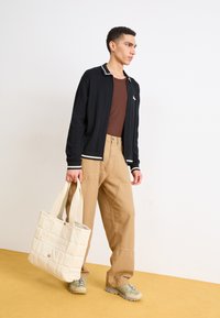 Czarna, żebrowana kurtka zapinana na zamek z białymi akcentami, brązowa koszula, beżowe spodnie cargo i pikowana torba w kolorze beżu w minimalstycznym otoczeniu.