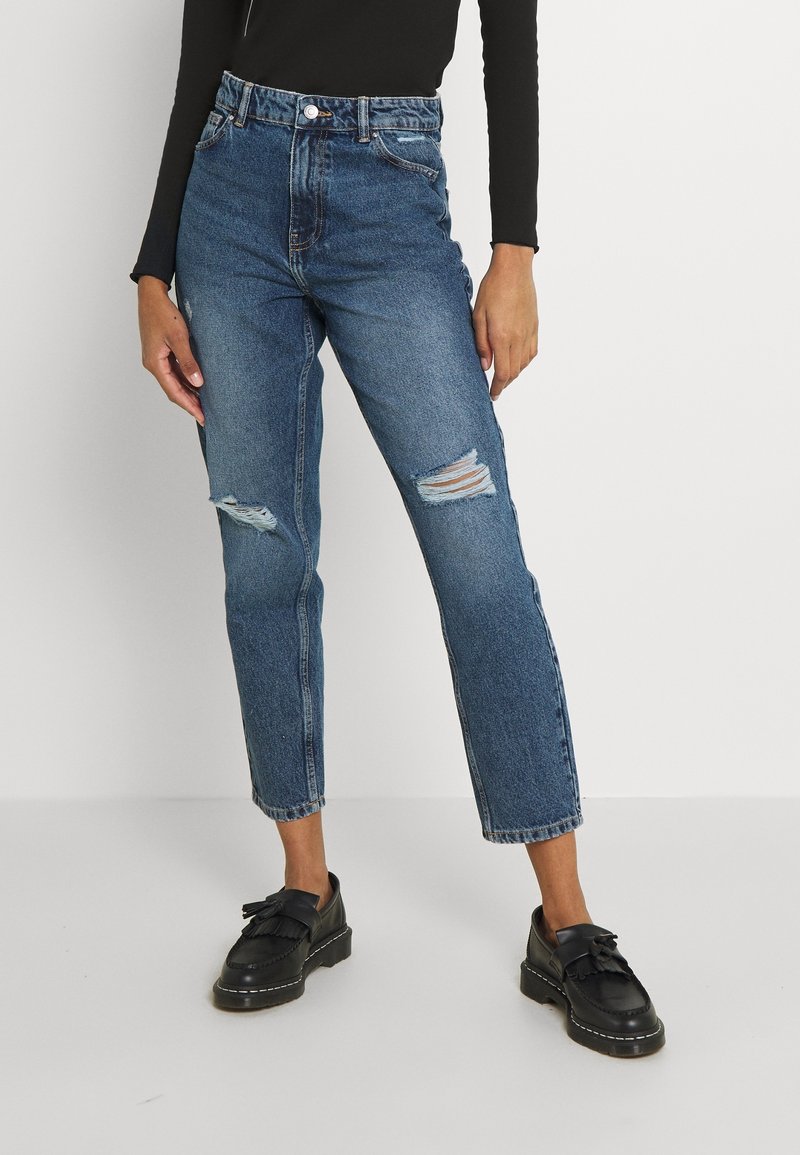 Högmidjade blå denimjeans med rivna knän och rak passform. Bärs tillsammans med svarta loafers med fransdetaljer.