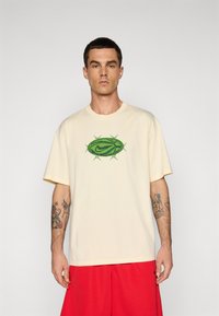 Mand med kort hår iført en cremet t-shirt med et grønt tornet ovalt Nike-logo og røde shorts, stående foran en ensfarvet lys baggrund.