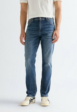 Homme portant un jean droit de couleur bleu moyen, un t-shirt blanc et des baskets blanches avec des bandes noires, debout contre un fond blanc uni.