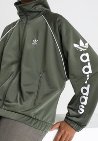 Olivgrön zip-up jacka med hög krage, vita detaljer och stor vit adidas-logga på högra ärmen. Slät, glänsande tyg.