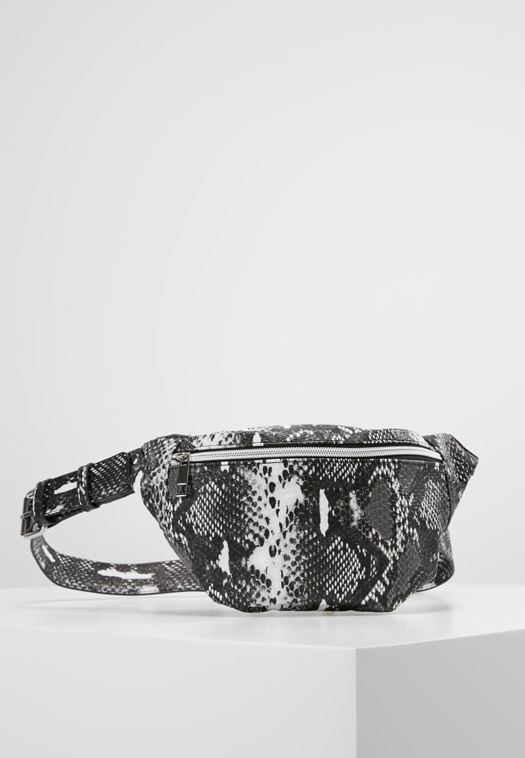 Sac banane à motif peau de serpent en noir et blanc avec fermeture éclair avant et sangle réglable, posé sur une surface blanche contre un fond uni.