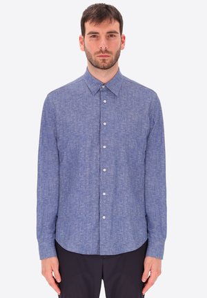 Uomo che indossa una camicia blu a maniche lunghe con trama e colletto appuntito, in piedi davanti a uno sfondo bianco.