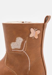 Botín de cuero marrón hasta el tobillo con un corazón de purpurina texturizado y una aplicación de mariposa brillante, con un forro suave y costuras decorativas.