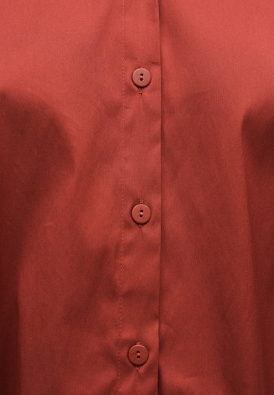 Tissu de chemise couleur rouille avec trois boutons ronds alignés verticalement et des coutures visibles le long de la patte de boutonnage.