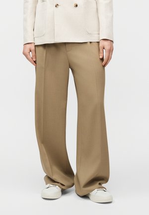 Oseba, oblečena v bež hlače širokih nogavic, bele superge in svetlo barvno blazerico, stoji pred enobarvnim ozadjem.