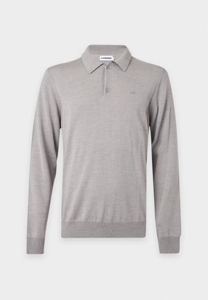 Mann trägt hellgraues, langärmeliges Poloshirt, dunkelblaue Hose, schwarze Schuhe, steht mit einer Hand in der Tasche vor einfachem weißem Hintergrund.
