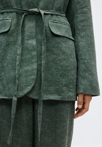 Tmavozelené ľanové sako s opaskom na uväzovanie, klopovými vreckami a zodpovedajúcimi nohavicami, zobrazené na dolnej časti trupu a ruke osoby.